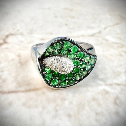 A close-up of a white gold ring featuring a calla lily-shaped setting filled with round green tsavorite gemstones and a central cluster of white diamonds, shown resting on a white background.

