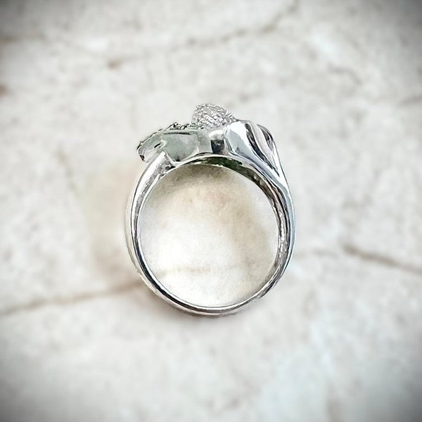 A close-up of the side profile of a white gold ring featuring a calla lily-shaped setting filled with round green tsavorite gemstones and a central cluster of white diamonds, shown resting on a white background.

