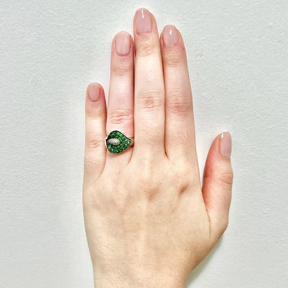 A close-up of a hand wearing a green tsavorite calla lily ring with small central clear diamond stones, set against a plain light gray background.

