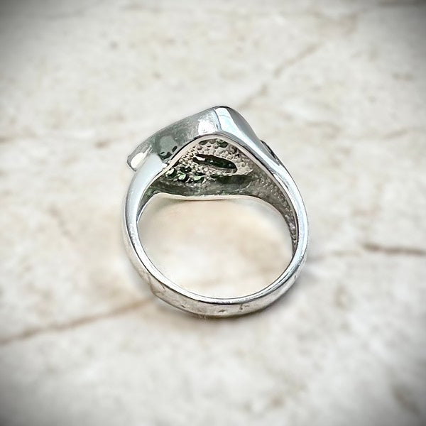 A close-up of the textured underside of a white gold ring featuring a calla lily-shaped setting filled with round green tsavorite gemstones and a central cluster of white diamonds, shown resting on a white knitted fabric.