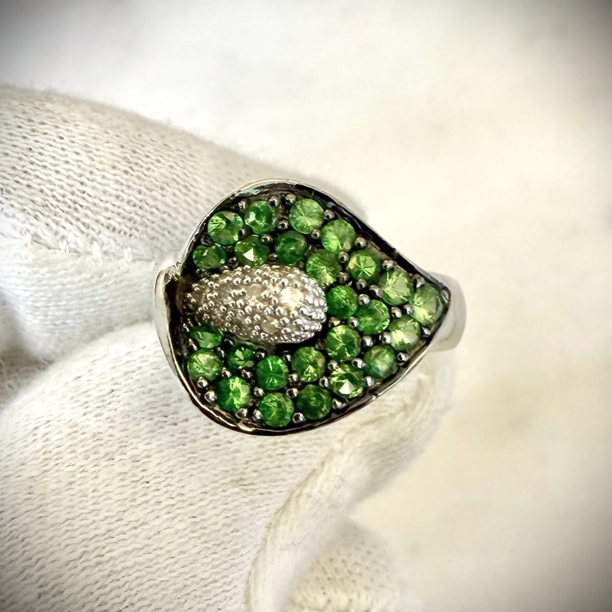 A close-up of a white gold ring featuring a calla lily-shaped setting filled with round green tsavorite gemstones and a central cluster of white diamonds, shown resting on a white knitted fabric.

