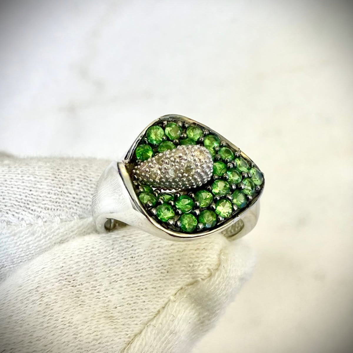 A close-up of a white gold ring featuring a calla lily-shaped setting filled with round green tsavorite gemstones and a central cluster of white diamonds, shown resting on a white knitted fabric.

