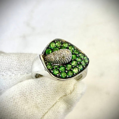 A close-up of a white gold ring featuring a calla lily-shaped setting filled with round green tsavorite gemstones and a central cluster of white diamonds, shown resting on a white knitted fabric.

