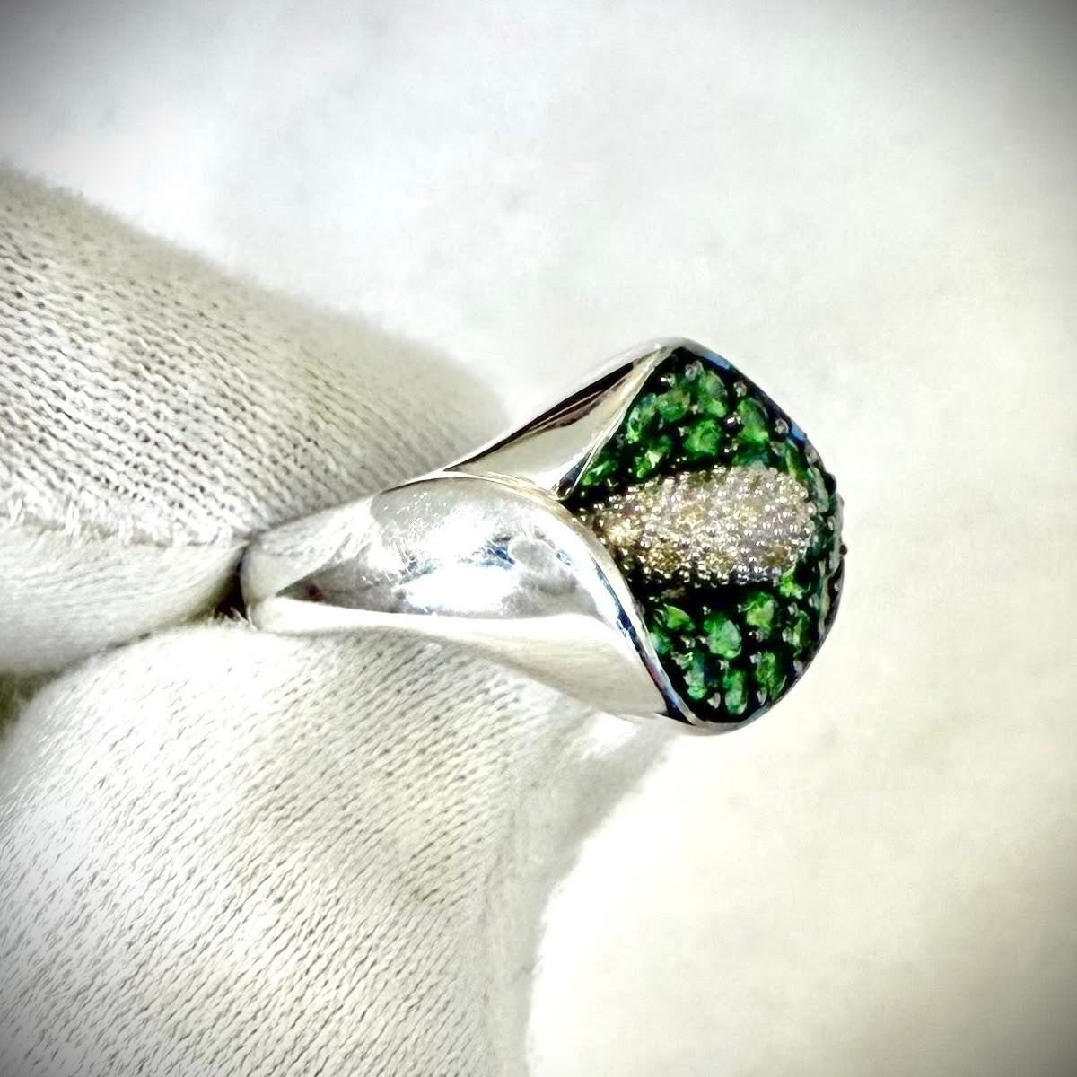 A close-up of a white gold ring featuring a calla lily-shaped setting filled with round green tsavorite gemstones and a central cluster of white diamonds, shown resting on a white knitted fabric.
