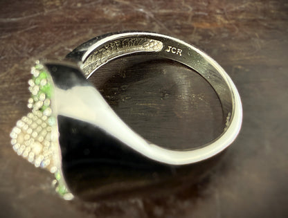 A close-up of the JCR maker's mark of a white gold ring featuring a calla lily-shaped setting filled with round green tsavorite gemstones and a central cluster of white diamonds, shown resting on a dark background.

