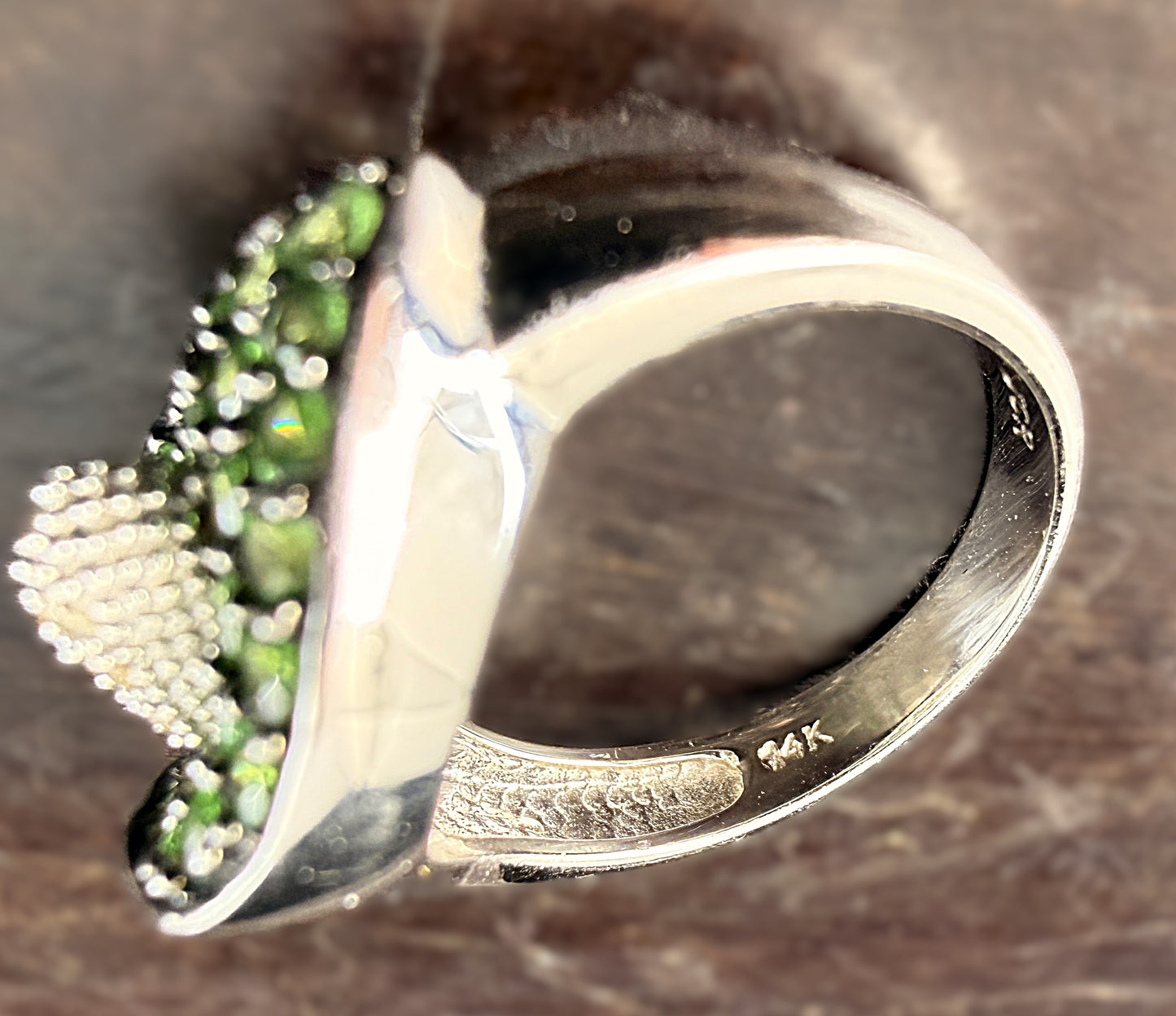 A close-up of the 14k hallmark of white gold ring featuring a calla lily-shaped setting filled with round green tsavorite gemstones and a central cluster of white diamonds, shown resting on a dark background.
