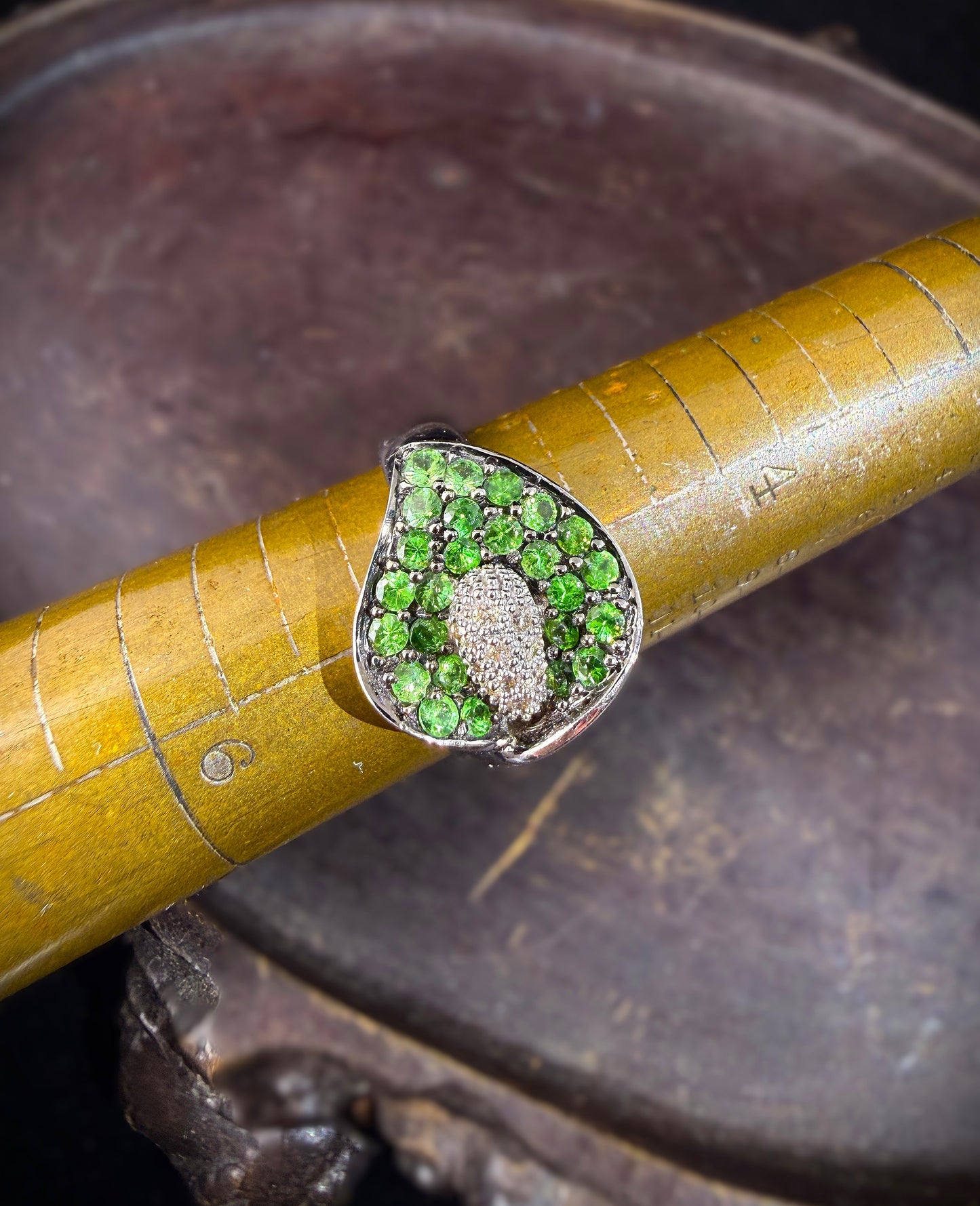 A close-up of a white gold ring featuring a calla lily-shaped setting filled with round green tsavorite gemstones and a central cluster of white diamonds, shown resting on an antique brass mandrel.

