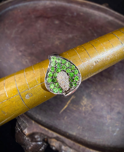 A close-up of a white gold ring featuring a calla lily-shaped setting filled with round green tsavorite gemstones and a central cluster of white diamonds, shown resting on an antique brass mandrel.

