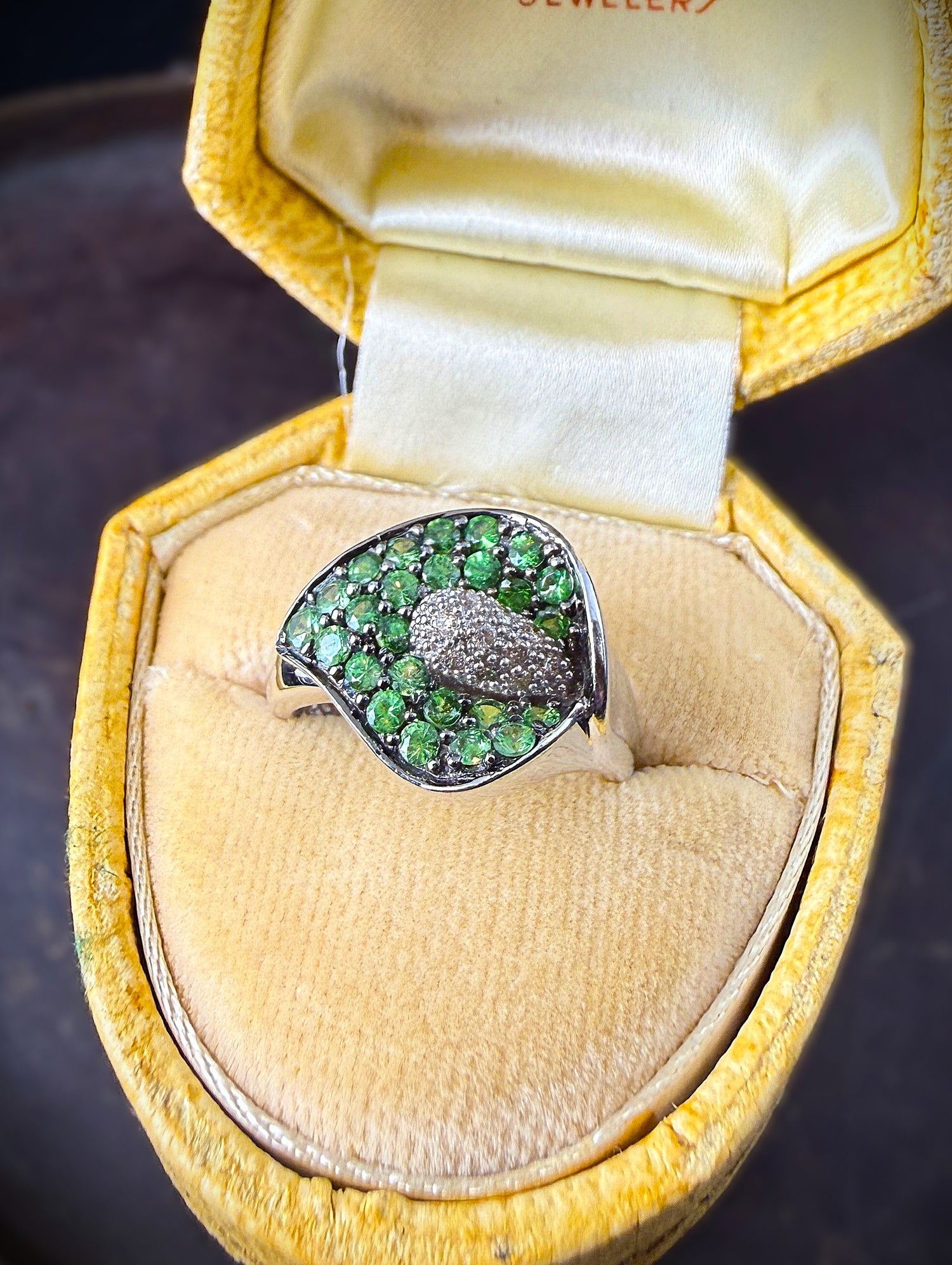 A white gold ring with a calla lily-shaped setting filled with green tsavorite garnet gems, displayed inside an open yellow velvet jewelry box.
